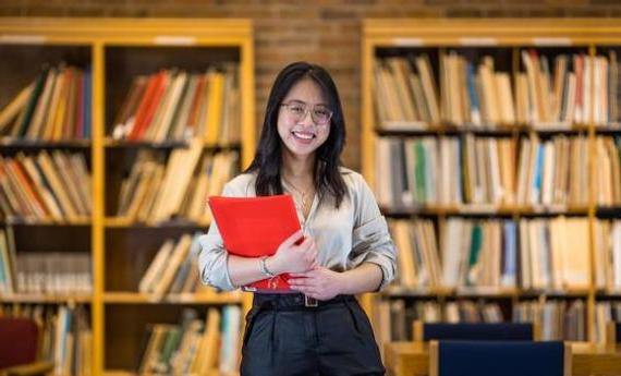 一个体育博彩app的学生站在图书馆里，手里拿着一个文件夹，面带微笑